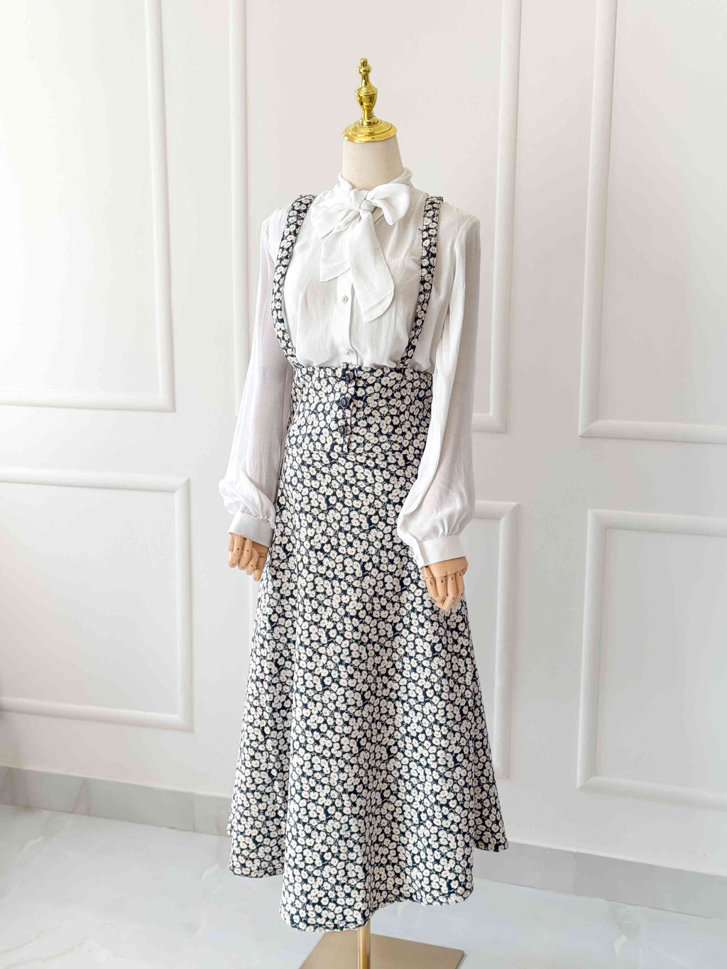 Mannequin wearing a white blouse with a large bow and a black floral dress against a white paneled wall.